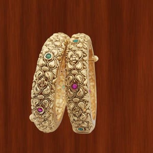 May include: Two gold bangles with detailed floral patterns and embedded gemstones. The bangles feature small, round green and pink gemstones. The design includes embossed floral motifs.