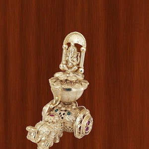 Pode incluir: Uma peça decorativa dourada com uma figura de divindade sentada dentro de uma moldura arqueada. A peça é intrincadamente detalhada com padrões florais e geométricos e adornada com pequenas pedras preciosas coloridas. O design inclui uma estrutura semelhante a um elefante.