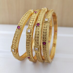 Peut inclure: Quatre bracelets dorés ornés de motifs complexes et de pierres précieuses. Chaque bracelet présente un motif répétitif avec de petites pierres rouges rondes et des pierres carrées transparentes. Le texte "4 pcs Bangles" est en bas.