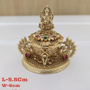 Puede incluir: Una caja de metal dorada con un diseño decorativo que presenta pavos reales y una pequeña figura de Ganesha en la parte superior. La caja tiene una tapa con un diseño de celosía y está adornada con piedras preciosas rojas y verdes. L-5,5 cm A-6 cm P-48 g