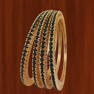 May include: A collection of gold-coloured bangles, each adorned with a line of teal gemstones. The bangles are arranged together, highlighting their detailed design and the contrast between the gold and teal colours. The background is a warm brown.