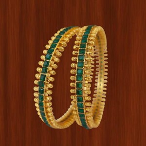 Peut inclure: Deux bracelets dorés ornés de pierres carrées vert émeraude. Les bracelets présentent un motif texturé avec de petits accents dorés ronds. Le fond est une surface en bois brun chaud.