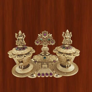 Puede incluir: Un artefacto religioso de color dorado con detalles intrincados. La pieza presenta dos recipientes ornamentados con figuras en la parte superior, un elemento decorativo central y una base adornada con piedras preciosas de colores. El fondo es marrón cálido.