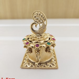Puede incluir: Una caja de metal dorada con un diseño de pavo real en la parte superior. La caja está decorada con piedras preciosas rojas y verdes y tiene una base cuadrada. L-5cm wt-30gm