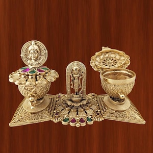 Può includere: Set di manufatti religiosi dorati e ornati. Il set include una figura centrale della divinità, due ciotole decorative e una struttura a forma di lampada, il tutto adornato con motivi intricati e pietre preziose colorate. Oggetti su una superficie di legno.