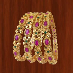 Peut inclure: Ensemble de bracelets dorés ornés de pierres précieuses ovales de couleur magenta. Les bracelets sont décorés de petites pierres vertes et de motifs floraux. Le fond est de couleur marron chaud.