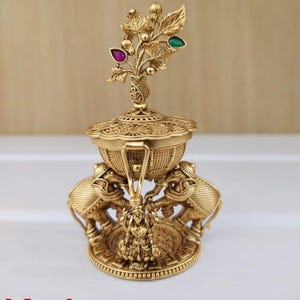 Puede incluir: Una caja de kumkum de color dorado con una tapa decorativa que presenta un diseño floral con piedras preciosas verdes y rosas. La caja está sostenida por dos elefantes y se asienta sobre una base circular con detalles intrincados. La caja mide aproximadamente 8 cm de largo.