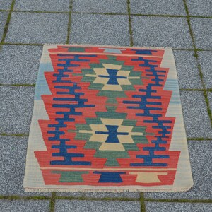 May include: A colorful rug with a geometric pattern in shades of red, blue, green, and beige. The rug has a fringed edge and is laid out on a gray tiled surface.