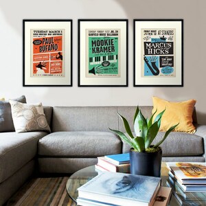 Op de afbeelding: Drie vintage concertposters in zwarte lijsten, elk met een andere muzikant. De eerste poster is rood, oranje en wit met de tekst "TUESDAY MARCH 1 RIVER HUNTINGTON PAUL BUFANO". De tweede poster is groen, blauw en wit met de tekst "SUNDAY FUNDAY FEST JUL. 24 GARFIELD HOUSE BALLROOM MOOKIE KRAMER and the Bi". De derde poster is blauw, geel en wit met de tekst "FRIDAY NIGHT JUNE 12 AT STANZOS MARCUS HICKS".