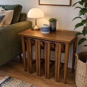May include: A set of three nested wooden tables with a warm brown finish, featuring tapered legs and a rectangular top. The tables are stacked together, creating a compact unit with a smooth surface that holds a white mushroom lamp and two vases.