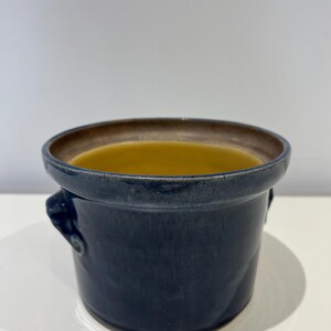 May include: A dark blue ceramic crock with a brown rim and two small handles. The interior is filled with a yellow liquid. The crock is on a white surface.