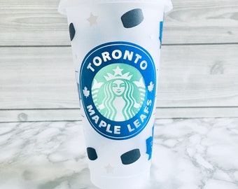 May include: A translucent plastic cold cup with a lid and straw, featuring a central circular logo with the text TORONTO MAPLE LEAFS in blue and a green siren graphic. The cup is decorated with small dark blue hockey pucks and white stars.