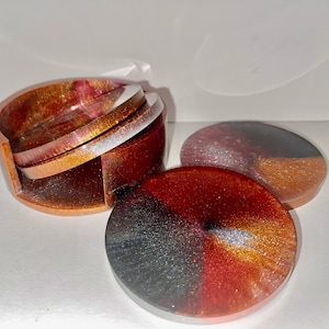 Puede incluir: Conjunto de cuatro posavasos redondos con un acabado brillante. Los posavasos están apilados en una pila con un soporte. Los posavasos tienen un degradado de colores negro, rojo y dorado.
