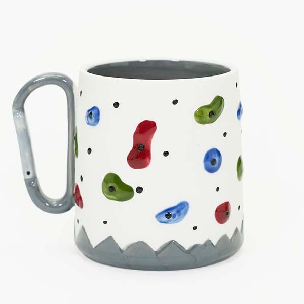 May include: A white ceramic mug with a grey handle and base, decorated with colourful climbing holds. The mug features red, green, and blue shapes, resembling climbing holds. The handle is designed to look like a carabiner.