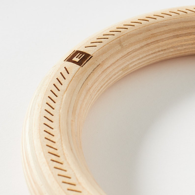 Puede incluir: Primer plano de un aro de gimnasia de madera con un patr&oacute;n de veta de madera y un peque&ntilde;o logotipo "E" grabado en el lateral. El aro est&aacute; hecho de madera natural y tiene un acabado liso y pulido.