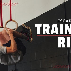 Puede incluir: Una persona haciendo un ejercicio de dominadas en anillos de gimnasia de madera colgados de correas rojas. Los anillos est&aacute;n suspendidos de un techo en un gimnasio. El texto "ESCAPE CLIMBING TRAINING RINGS" se muestra en letras blancas sobre una pared de ladrillo negro.