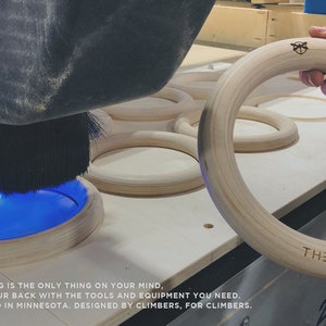 Puede incluir: Primer plano de un anillo de gimnasia de madera que se est&aacute; fabricando en una m&aacute;quina CNC. El anillo es de color marr&oacute;n claro y tiene el texto "THE WILDERNESS" grabado.