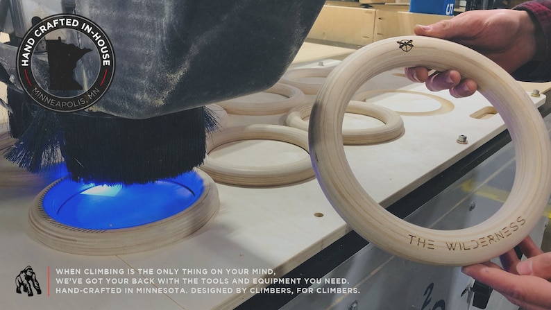 Puede incluir: Primer plano de un anillo de gimnasia de madera que se est&aacute; fabricando en una m&aacute;quina CNC. El anillo es de color marr&oacute;n claro y tiene el texto "THE WILDERNESS" grabado.