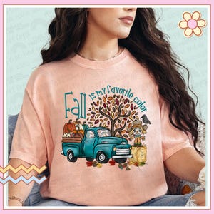 May include: A pink t-shirt featuring a graphic design of a blue pickup truck carrying pumpkins and a scarecrow. The text "Fall is my favourite colour" is written in a teal font.