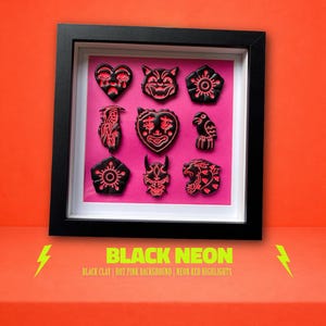 Puede incluir: Una obra de arte enmarcada con nueve diseños de arcilla negra con detalles en rojo neón sobre un fondo rosa. Los diseños incluyen un corazón, un gato, una flor, una parca, una cara sonriente, un pájaro, una flor, un demonio y un tigre. El marco es negro y el fondo es naranja. Texto: "BLACK NEON".