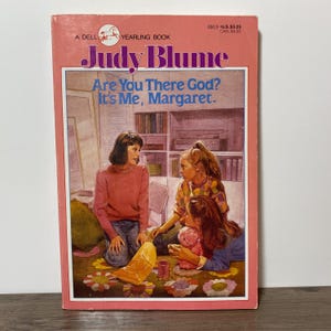 Peut inclure: Un livre de poche vintage intitulé "Are You There God? It's Me, Margaret." de Judy Blume. La couverture présente une illustration de trois filles dans une pièce. Le livre a une couverture rose avec le texte "A Dell Yearling Book."