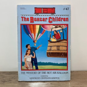May include: A vintage paperback book titled "The Boxcar Children: The Mystery of the Hot Air Balloon." The cover features a colorful hot air balloon, a family, and a red train car illustration against a blue sky. The author is Gertrude Chandler Warner.