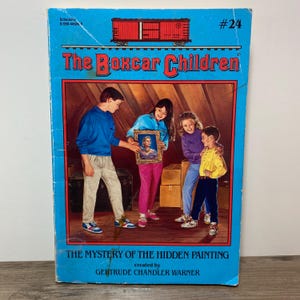 May include: A vintage copy of "The Boxcar Children: The Mystery of the Hidden Painting" book. The cover features a blue background with the title and a red boxcar illustration. Four children are shown examining a framed painting.