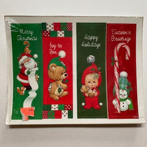 May include: Four Christmas-themed gift tags with red, green, and white colors. The tags feature illustrations of Santa Claus, a teddy bear, a baby, and a snowman. The tags have the text "Merry Christmas", "Joy to You", "Happy Holidays", and "Season's Greetings".