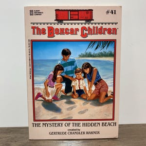 May include: A vintage paperback book titled "The Boxcar Children" with a beach scene illustration. The cover shows four children on a sandy beach. The title is "The Mystery of the Hidden Beach" by Gertrude Chandler Warner.