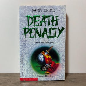 May include: A paperback book titled "Death Penalty" by Dennis Hamley. The cover features a soccer ball with a dagger, set against a green and gray background. The text "Point Crime" is at the top.