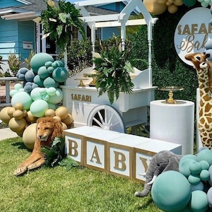 May include: A white baby carriage with a gold sign that says "Safari" and a white pedestal with a gold plate on top. There are green and gold balloons, a stuffed lion, a stuffed elephant, and wooden blocks that spell out "BABY".