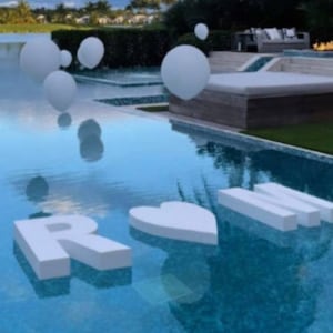 May include: White letters spelling "ROM" float in a blue swimming pool. A heart-shaped letter "O" is in the middle. White balloons are floating in the background.