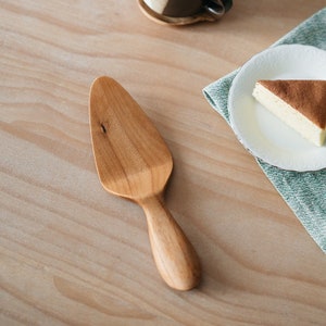 Könnte beinhalten: Ein hölzerner Kuchenschieber in hellbrauner Farbe mit einem glatten, abgerundeten Griff. Der Schieber befindet sich auf einer hellbraunen Holzoberfläche.