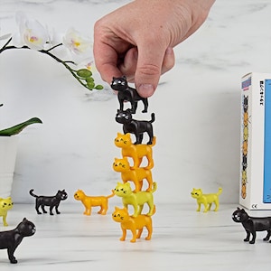 May include: A hand is seen stacking black and yellow cat figurines. The figurines are in various shades of yellow and black. Some figurines are stacked on top of each other, while others are scattered around. A box with Japanese text is in the background.