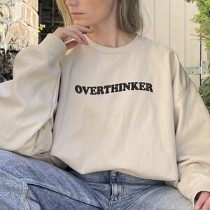 May include: A beige sweatshirt with the text "OVERTHINKER" embroidered in black on the front.