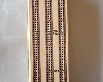 May include: A wooden cribbage board with a hinged lid. The board has three tracks with numbered holes for pegs, a start and finish area, and decorative etched details. The board is made of light-coloured wood.