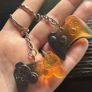 May include: Two keychains featuring controller designs. One keychain has a controller shape in black and orange, and the other has a controller shape in orange and black. Both keychains are attached to a silver chain.