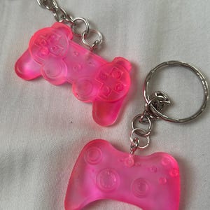 May include: Two vibrant pink keyrings in the shape of video game controllers. Each controller features detailed button and directional pad designs. The keyrings are attached to silver metal key rings and chains.
