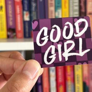 May include: A sticker with a purple and white checkered background. The words "GOOD GIRL" are written in white with a purple outline. There are small pink hearts in the corners.