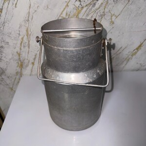 Peut inclure: Un bidon à lait cylindrique en métal argenté doté d'une large ouverture sur le dessus, d'un col effilé lisse et d'une poignée articulée robuste fixée sur les côtés. Le récipient présente un design simple et utilitaire avec une finition mate et une barre métallique.
