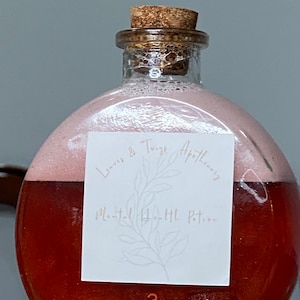 May include: A glass bottle with a cork stopper filled with a red liquid. The bottle has a white label with the text "Leaves & Twigs Apothecary" and "Moonlit Herbal Potion".