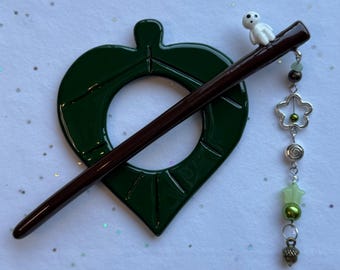 May include: A dark green leaf-shaped hair pin with a brown stick and a small white figurine. The pin has a dangling charm with green beads, a flower, and an acorn. The hair accessory is a unique and decorative item.