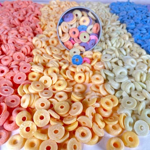 May include: A close-up of a variety of colorful cereal rings. The cereal is arranged in rows of different colors, including pink, yellow, and blue. A small bowl of the cereal is in the center of the image.