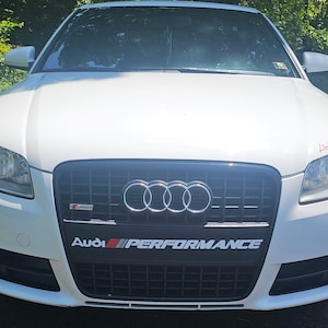 May include: White Audi sedan with a black grille and the words "Audi Performance" in red and black lettering.