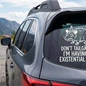 May include: A white vinyl car decal featuring a cartoon rat wearing a party hat. The text reads "DON'T TAILGATE ME I'M HAVING AN EXISTENTIAL CRISIS" on the rear window of a gray car, with mountains in the background.