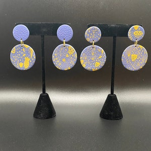 May include: Two pairs of handmade earrings. Each earring features a small, textured, round stud in periwinkle blue, connected to a larger, circular pendant. The pendants are periwinkle blue with a yellow floral pattern. The earrings are displayed on black stands.