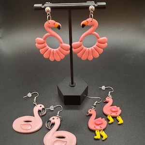 May include: Pink flamingo earrings in various designs. Two pairs feature a flamingo silhouette with a fan-shaped base. Two other pairs show a flamingo shape with a curved neck. The last pair has yellow boots.