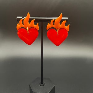 May include: A pair of red heart-shaped earrings with orange flames on top. The earrings are displayed on a black stand.