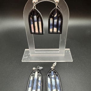 May include: A pair of black and white stained glass window earrings with a blue and white sky design. The earrings are on a clear display stand.