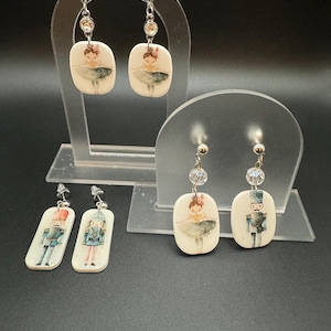 May include: Three pairs of dangle earrings featuring a ballerina and a nutcracker. The earrings are made of white ceramic with a painted design and have a crystal bead on the top of the dangle.